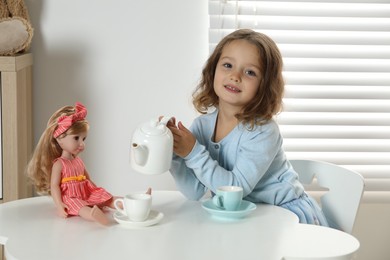 Photo of Cute little girl playing tea party with doll at white table in room