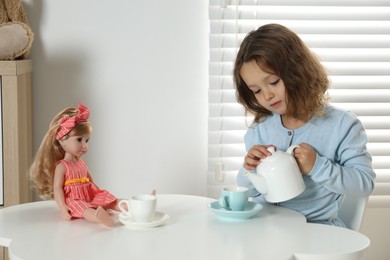 Photo of Cute little girl playing tea party with doll at white table in room