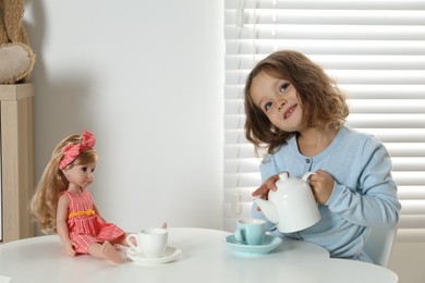 Photo of Cute little girl playing tea party with doll at white table in room