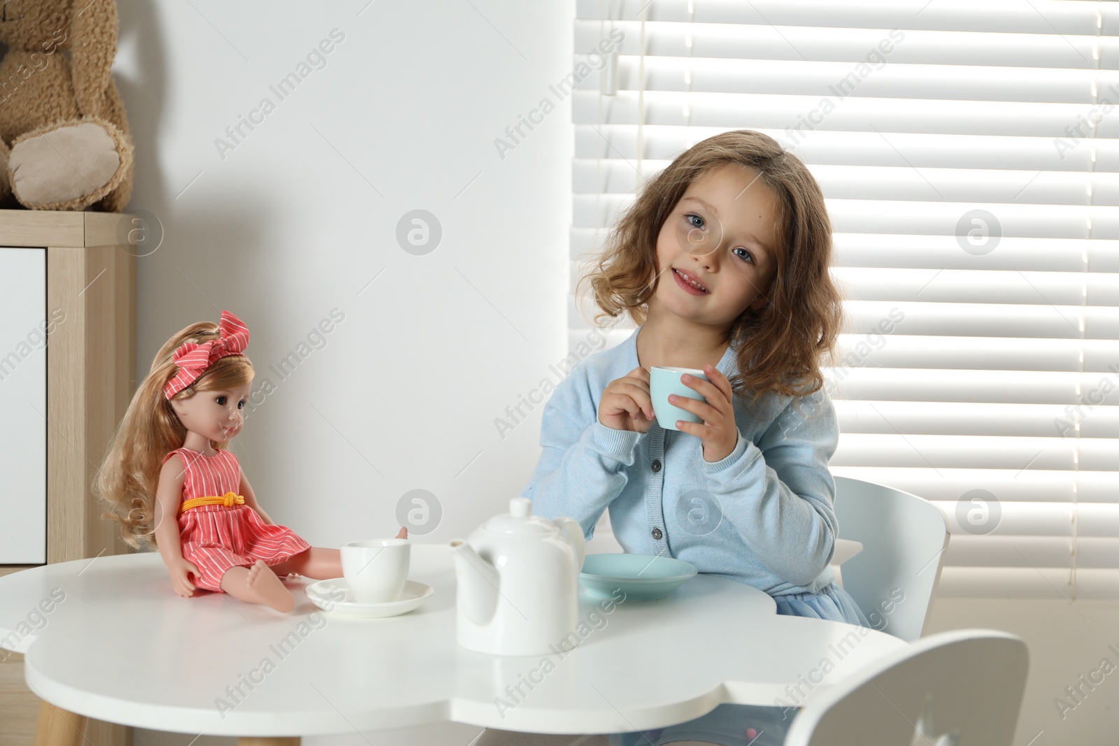 Photo of Cute little girl playing tea party with doll at white table in room