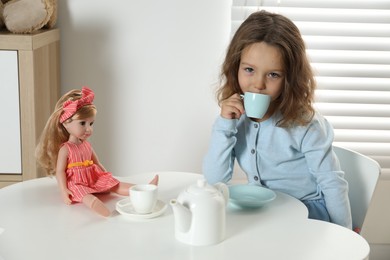 Photo of Cute little girl playing tea party with doll at white table in room