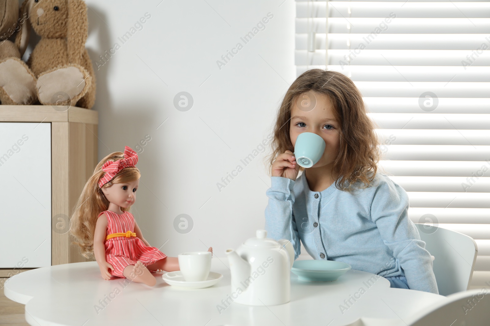 Photo of Cute little girl playing tea party with doll at white table in room