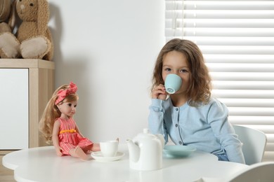 Photo of Cute little girl playing tea party with doll at white table in room