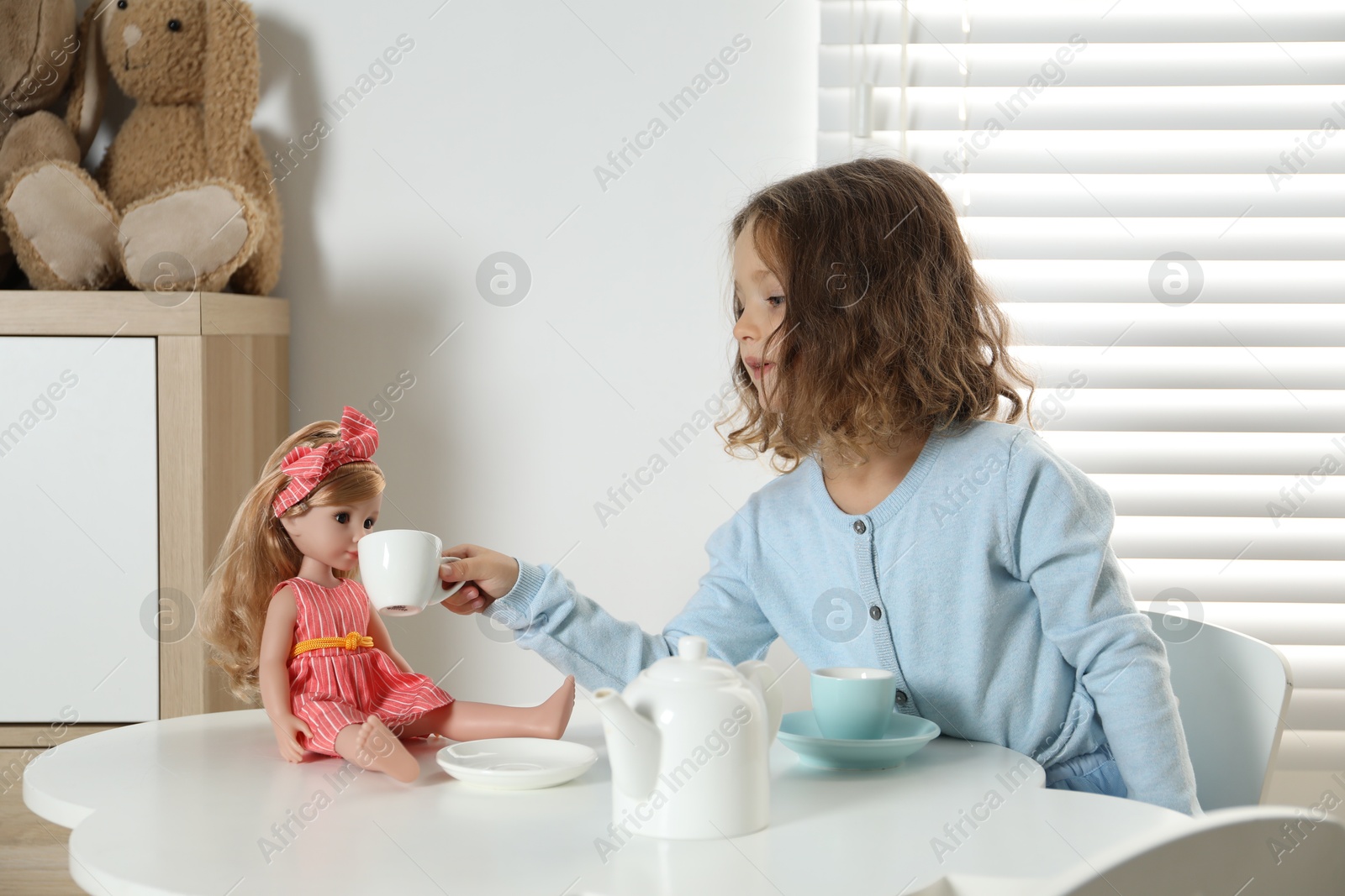 Photo of Cute little girl playing tea party with doll at white table in room