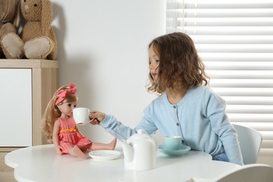 Photo of Cute little girl playing tea party with doll at white table in room