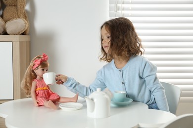 Photo of Cute little girl playing tea party with doll at white table in room