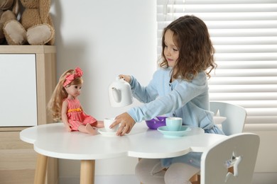 Photo of Cute little girl playing tea party with doll at white table in room