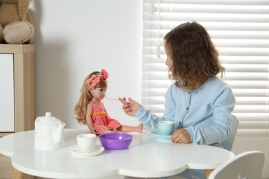 Photo of Little girl playing with doll and toy dishware at table in room