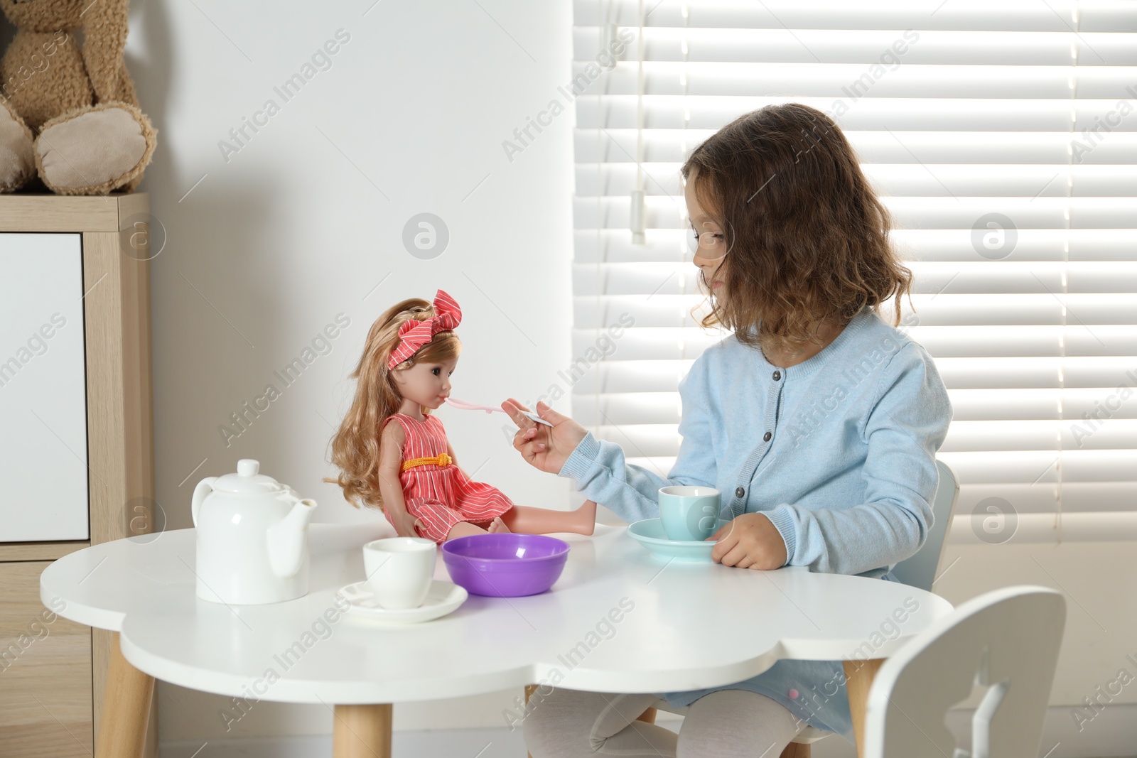 Photo of Little girl playing with doll and toy dishware at table in room