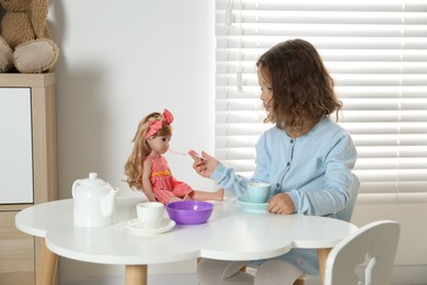 Photo of Little girl playing with doll and toy dishware at table in room