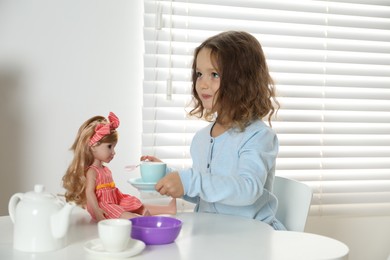 Photo of Cute little girl playing with doll and toy dishware at table in room