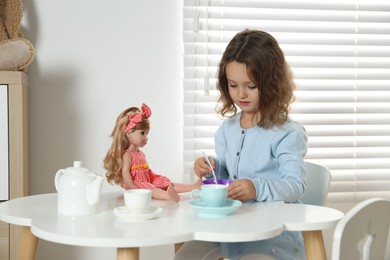 Photo of Cute little girl playing with doll and toy dishware at table in room