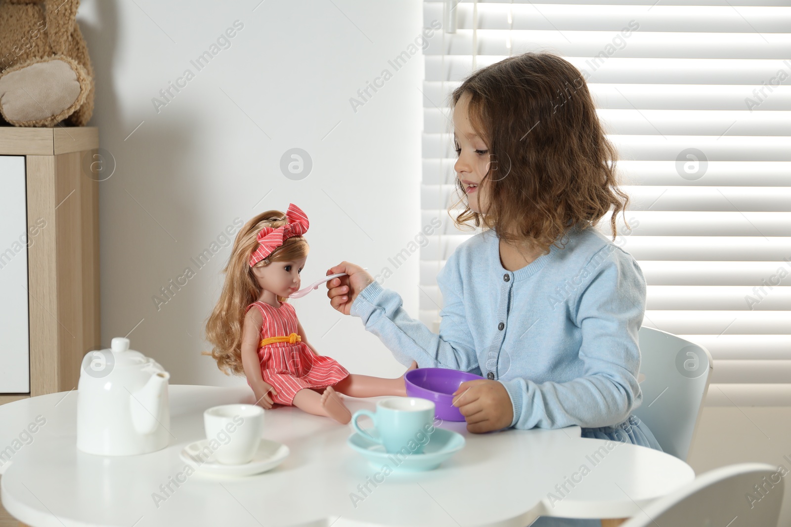 Photo of Cute little girl playing with doll and toy dishware at table in room