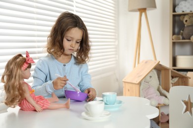 Photo of Cute little girl playing with doll and toy dishware at table in child's room