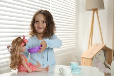 Photo of Cute little girl playing with doll and toy dishware at table in child's room