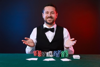 Professional croupier at table with playing cards and casino chips against color background