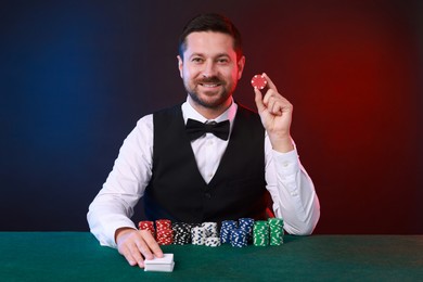 Professional croupier with playing cards and casino chips at gambling table against color background