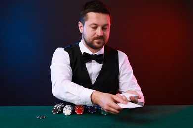 Professional croupier with playing cards at gambling table against color background