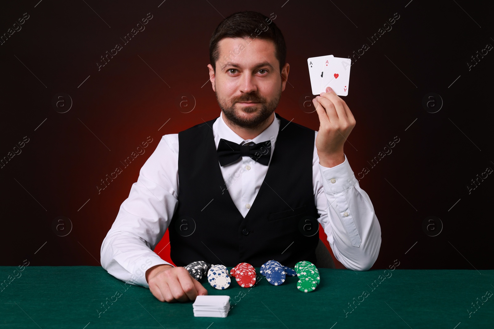 Photo of Professional croupier with playing cards at gambling table against dark red background