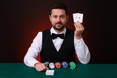 Photo of Professional croupier with playing cards at gambling table against dark red background