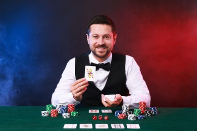 Professional croupier with playing cards at gambling table against color background with smoke