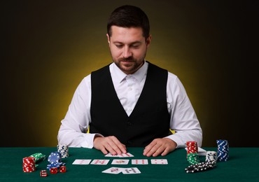 Professional croupier with playing cards at gambling table on dark yellow background