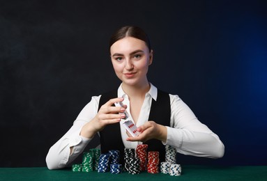 Professional croupier with chips shuffling playing cards at gambling table on color background