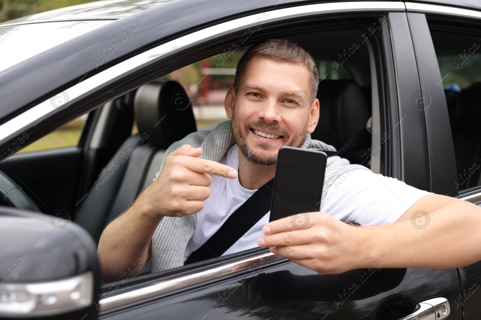 Photo of Happy man pointing at smartphone in modern car, view from outside