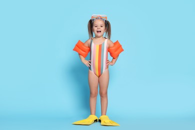 Cute little girl in swimsuit with swim armbands and fins on light blue background