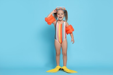 Cute little girl in swimsuit with swim armbands and fins on light blue background