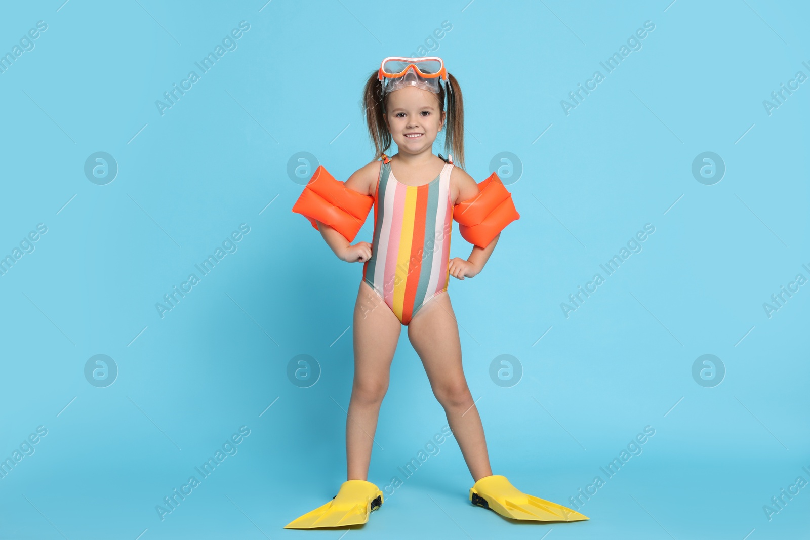 Photo of Cute little girl in swimsuit with swim armbands and fins on light blue background