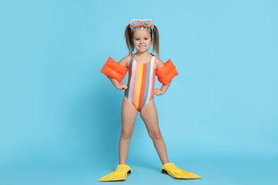 Cute little girl in swimsuit with swim armbands and fins on light blue background