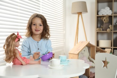 Photo of Cute little girl playing with doll and toy dishware at table in child's room