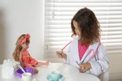 Photo of Little girl playing doctor with doll at table in room