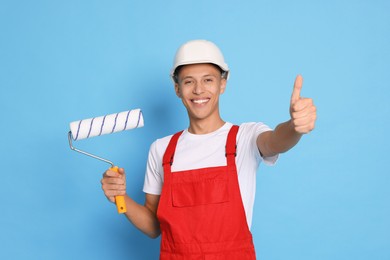 Photo of Portrait of young decorator with paint roller showing thumbs up on light blue background