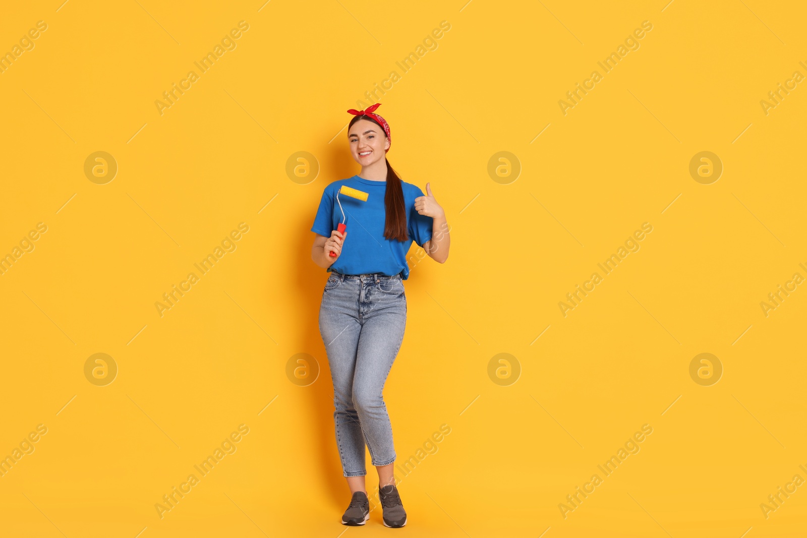 Photo of Young decorator with paint roller showing thumbs up on orange background