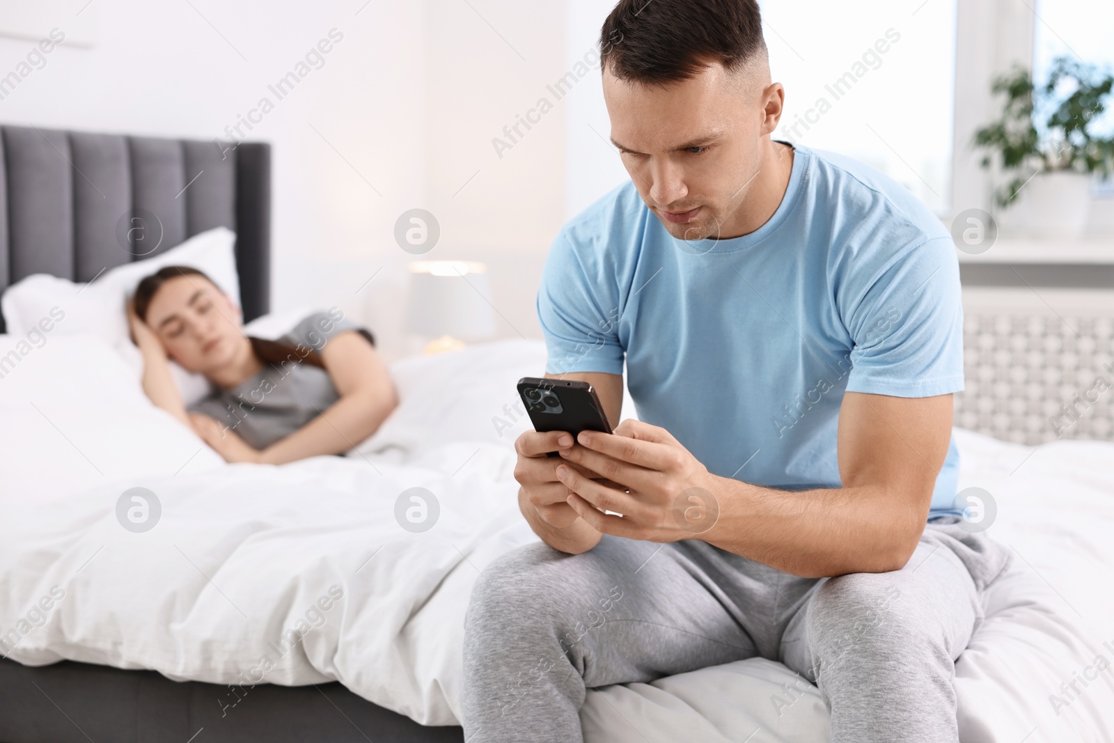 Photo of Jealous man checking wife's smartphone while she sleeping in bedroom, selective focus