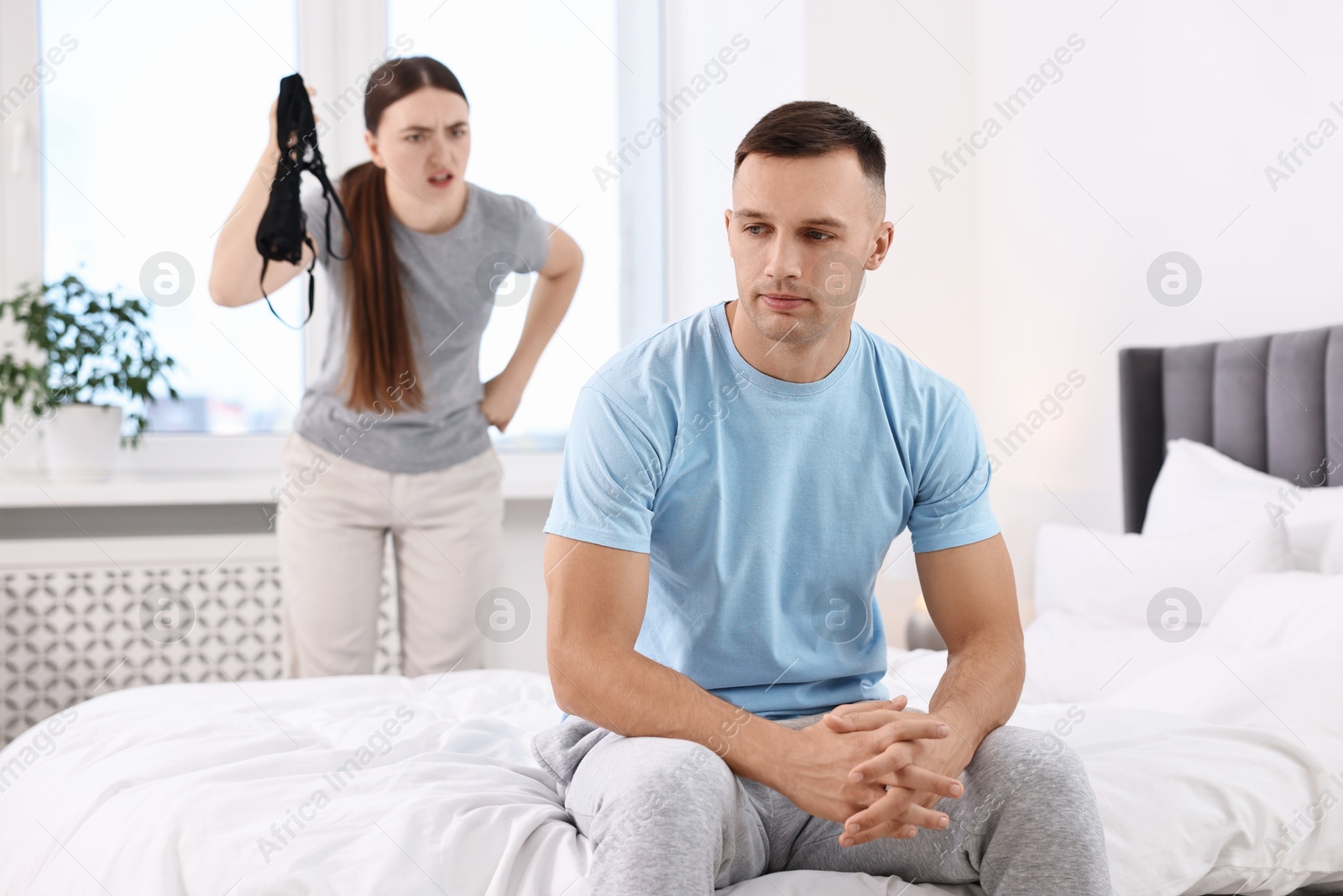 Photo of Angry wife showing mistress bra as proof to her unfaithful husband while he ignoring her in bedroom, selective focus