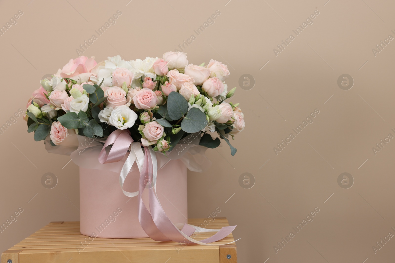 Photo of Gift box with bouquet of beautiful roses and eucalyptus branches on wooden nightstand against beige background. Space for text