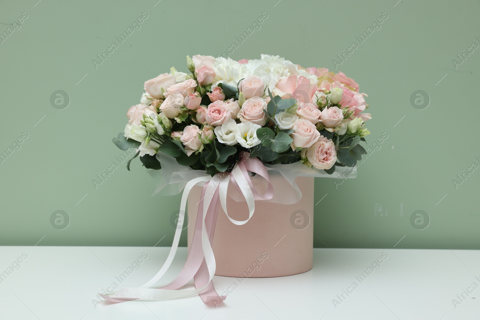 Photo of Gift box with bouquet of beautiful flowers and eucalyptus branches on white table against green background