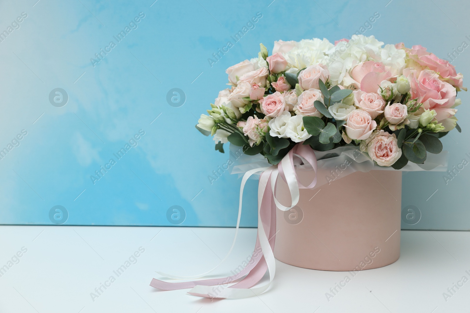 Photo of Gift box with bouquet of beautiful flowers and eucalyptus branches on white table against light blue background. Space for text