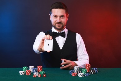 Photo of Professional croupier with playing cards at gambling table against color background