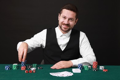 Photo of Professional croupier at gambling table with playing cards, casino chips and dice against black background