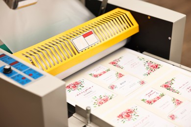 Photo of Modern roll laminator and paper sheet with invitation cards indoors, closeup. Printing house