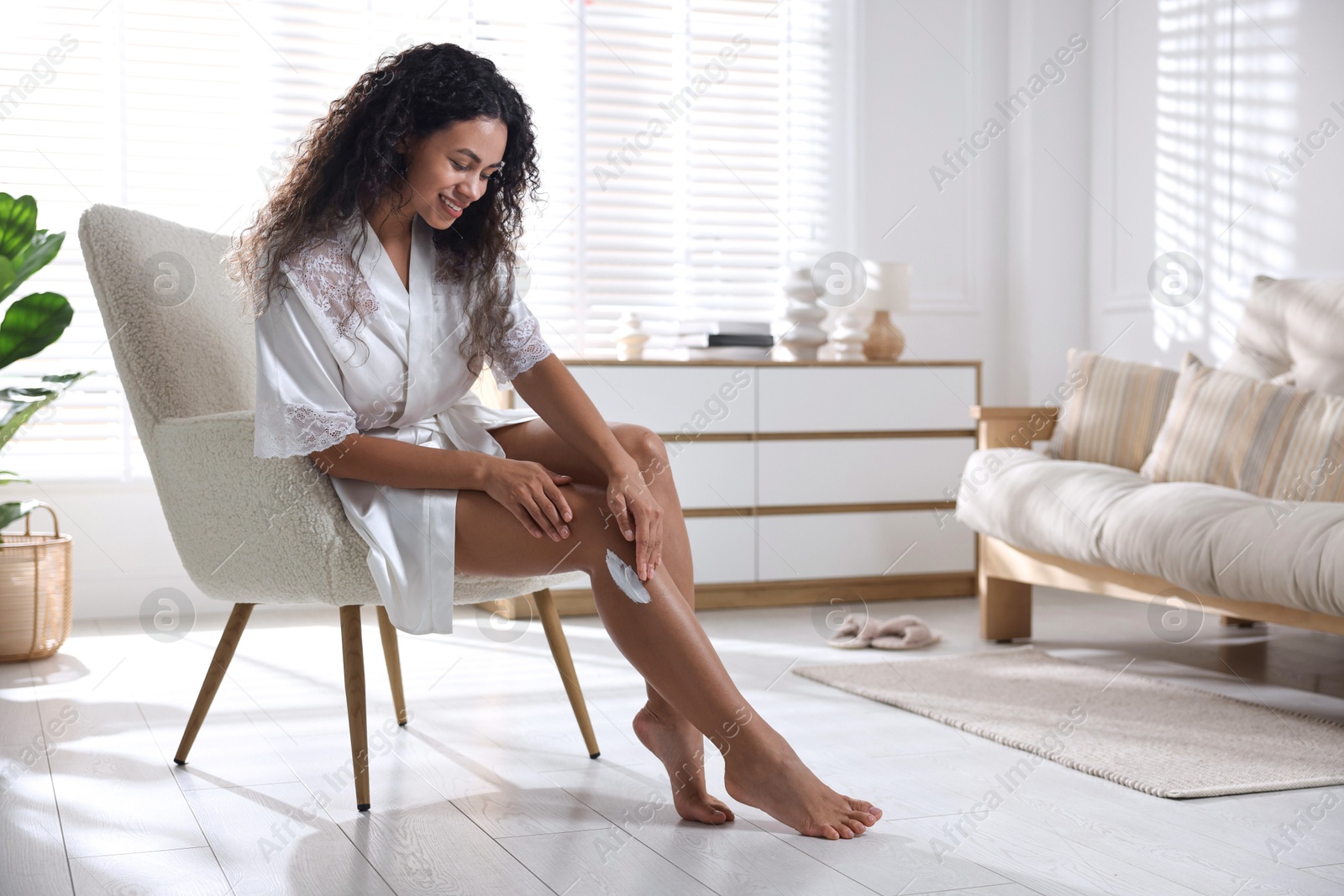 Photo of Young woman applying cream onto leg in armchair at home