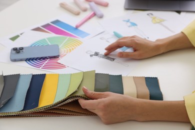 Photo of Fashion designer with sketch of outfit choosing fabric among colorful samples at white table in workshop, closeup