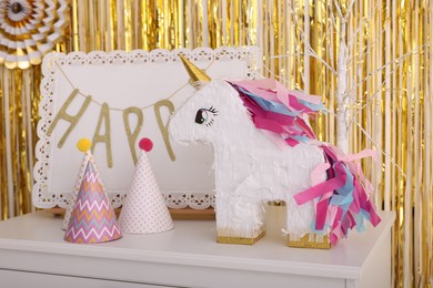 Photo of Bright pinata in shape of unicorn and party accessories on white chest of drawers against foil fringe curtain indoors