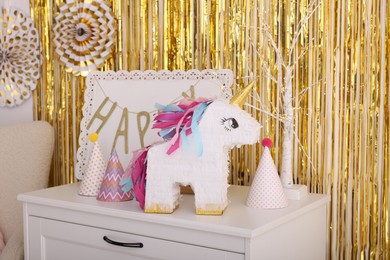 Photo of Bright pinata in shape of unicorn and party accessories on white chest of drawers against foil fringe curtain indoors