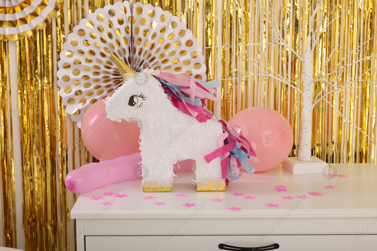 Photo of Bright pinata in shape of unicorn and party accessories on white chest of drawers against foil fringe curtain indoors