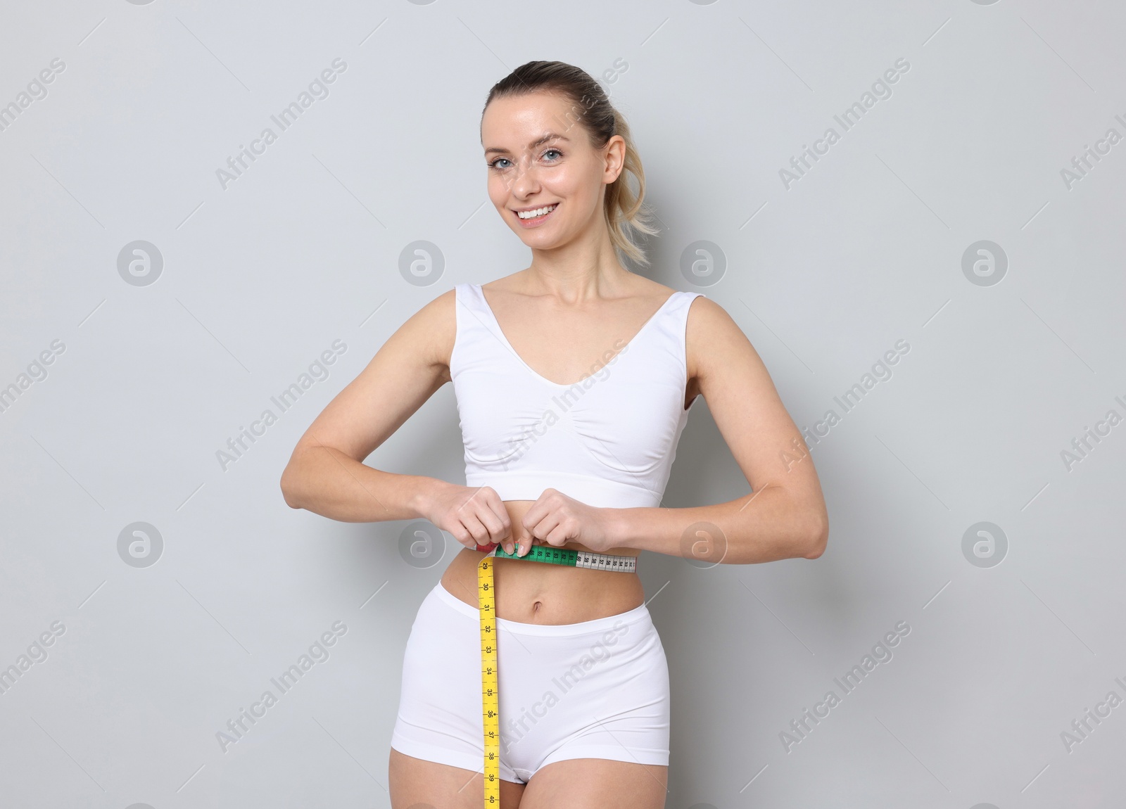 Photo of Weight loss. Happy woman measuring her slim waist with tape on light grey background